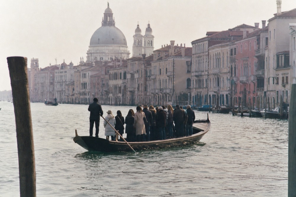 A flat-bottomed ferry gondola carrying standing commuters across a wide waterway, palazzi and a church dome behind
