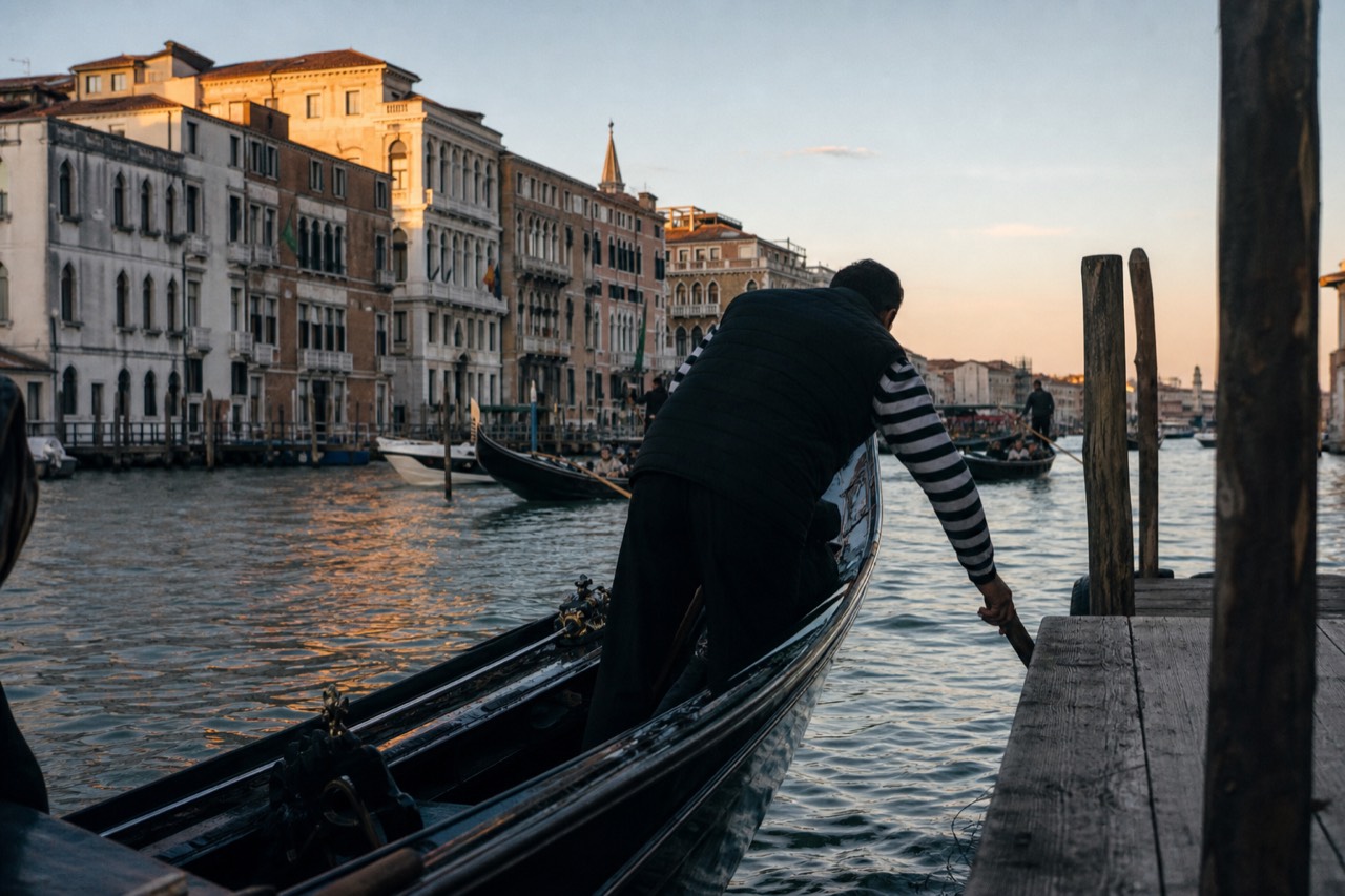 A gondolier's hand reaching down from the stern of a black boat toward a wooden dock, canal water reflecting late afternoon sun