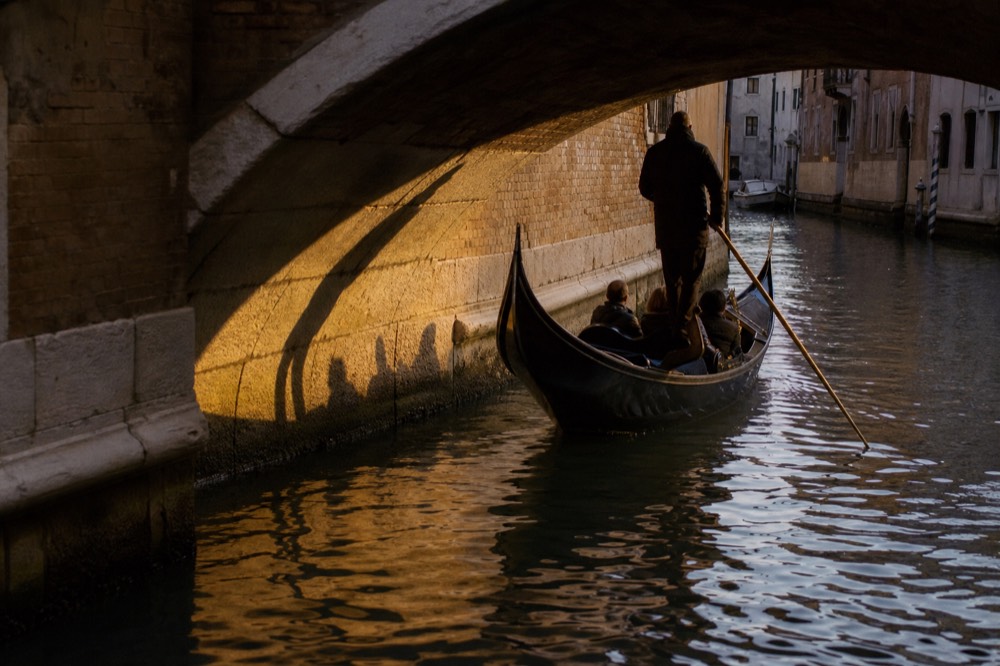 A rowed black boat passing under a low stone bridge in a narrow canal, the oarsman's shadow thrown against the wall