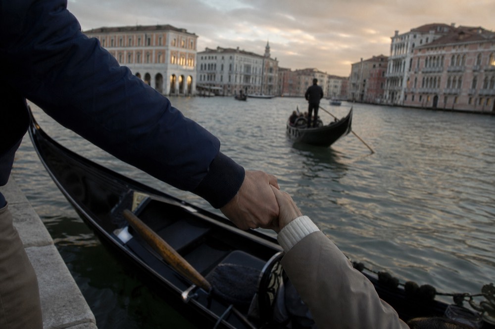 A passenger's hand gripping a gondolier's forearm while stepping down from a stone quay into a rocking boat, no faces in frame
