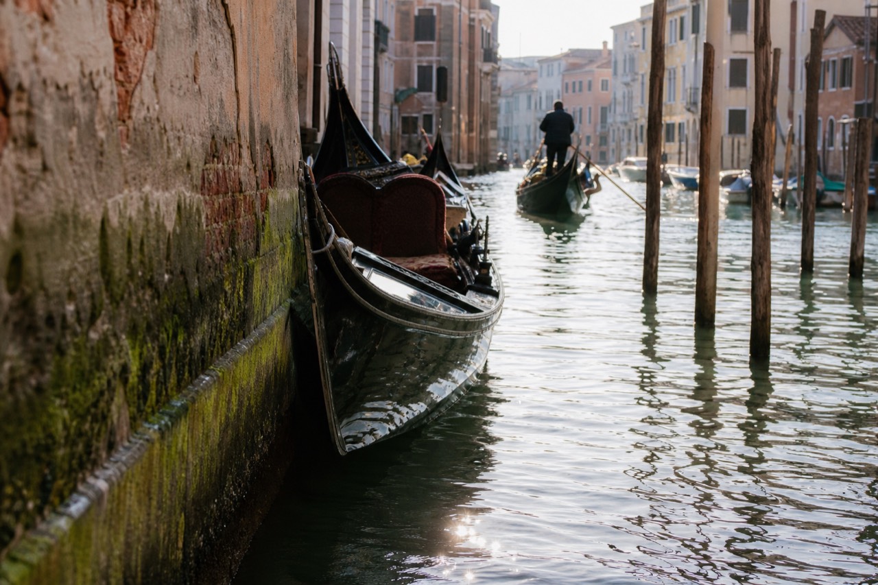 A gondola's long slender black hull seen from the waterline, showing how narrow the beam sits against a mossy canal wall