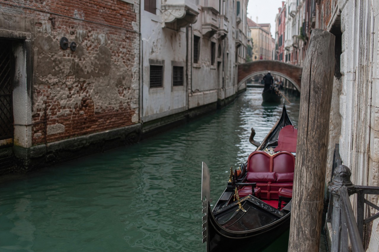A black gondola with a red velvet bench waiting at a wooden mooring post on a narrow Venetian canal in late morning light