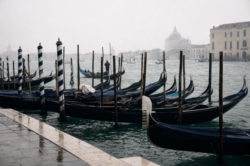 A row of black gondolas rocking at their striped mooring poles on a wide lagoon channel under an overcast sky