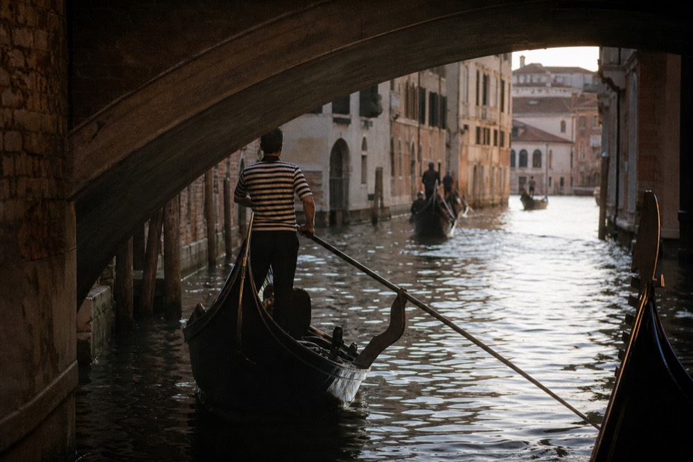 A gondolier in a striped shirt seen from behind at the stern oar as his boat enters a shaded canal beneath a low stone bridge