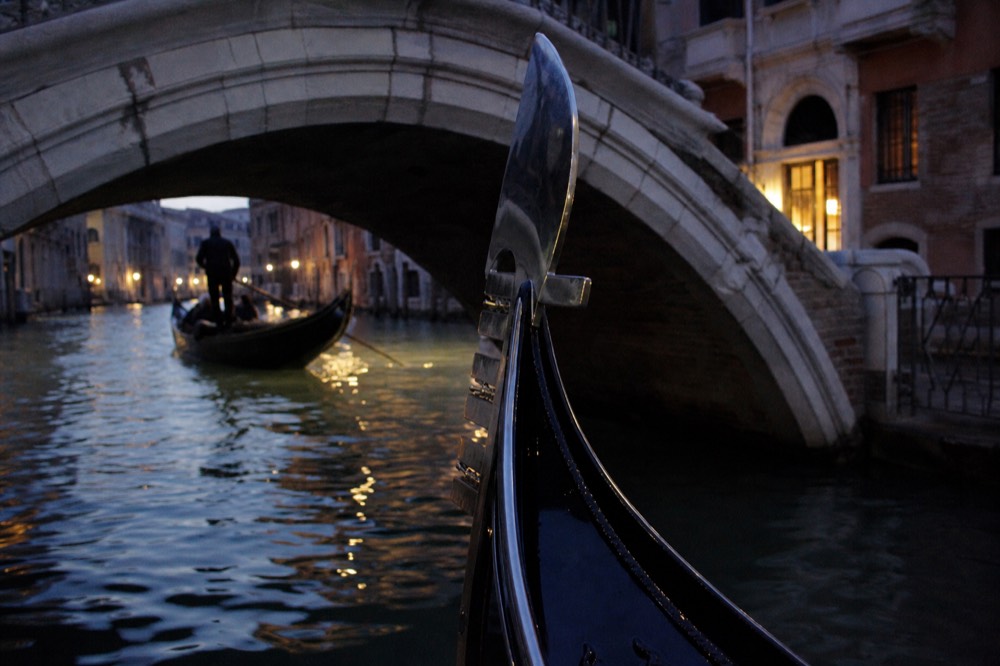 A gondola prow ornament in polished steel rising above dark water with a stone bridge arch framing the scene behind