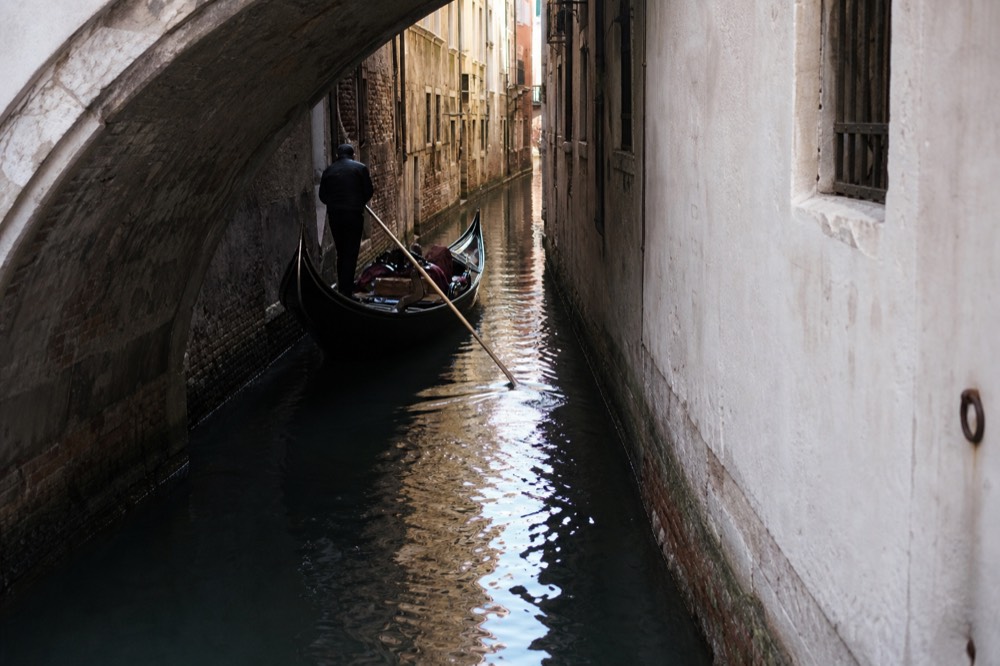A single gondola turning into a very narrow canal between tall plastered walls, the water almost black in the shade