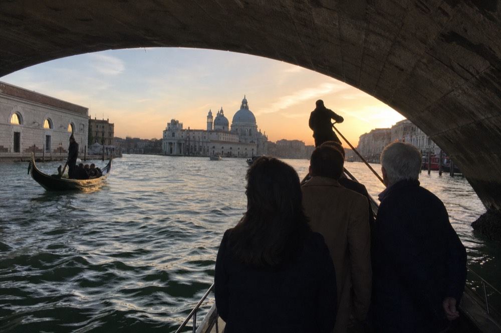 Commuters standing upright in a plain ferry gondola crossing wide green water, a domed church silhouetted behind them at dusk
