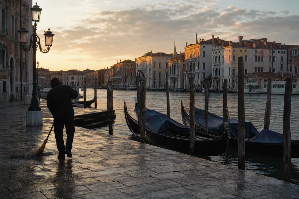 An empty morning quay with three gondolas waiting under covers and a single figure sweeping the stones in low sunlight