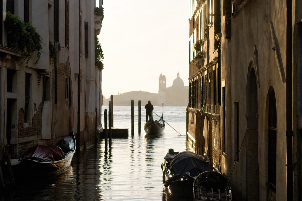 A gondolier in a striped shirt standing at the stern rowing with a long oar, seen in silhouette against pale reflected water at the end of a canal