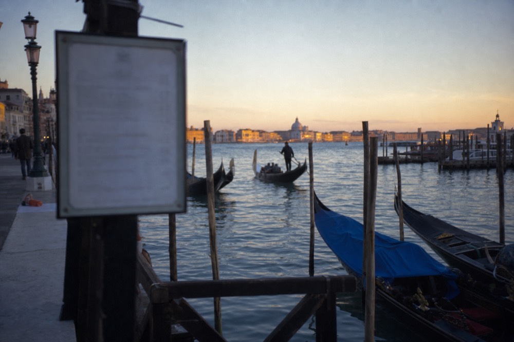 A printed tariff board mounted on a mooring post at a gondola landing, blurred beyond legibility, with moored boats and evening water behind