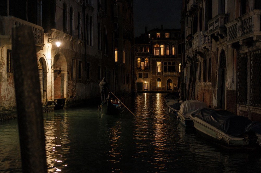 Night canal scene with strings of window light broken into long streaks on black water and a single lamp glowing above an arched doorway