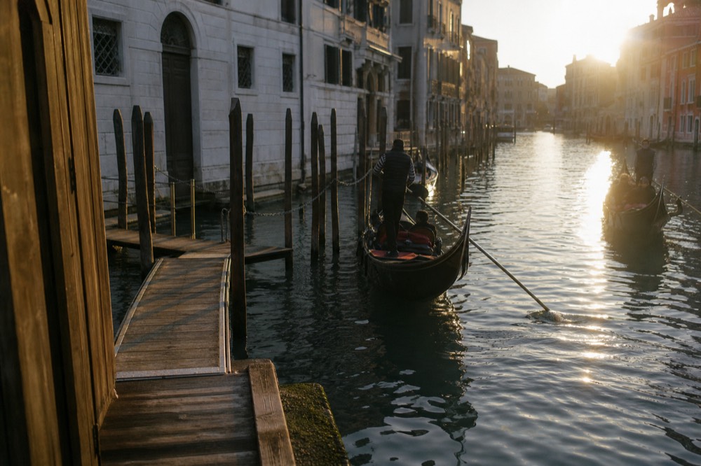 Wooden gondola pontoon with a low gangway and a rack of dark mooring posts on a small side canal, morning shadow across the water