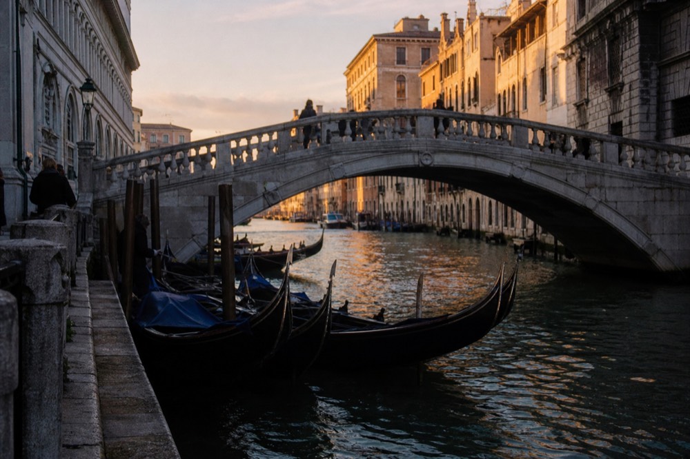 Gondolas gathered against a narrow quay directly beneath a wide stone bridge, sunlight catching the water between the hulls