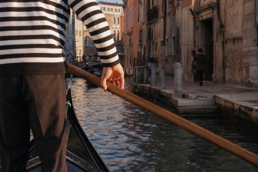Close view of a gondolier's striped shirt and oar hand at the stern rowing position, canal water and stone steps below