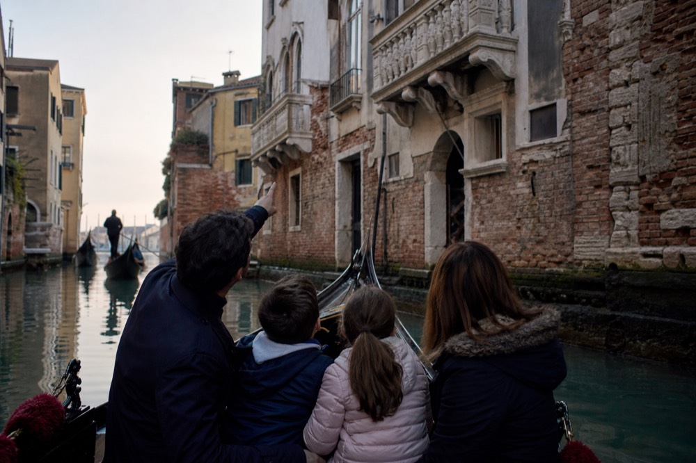 A family of four seated together in a gondola from behind, an adult pointing up at a carved stone balcony above the water